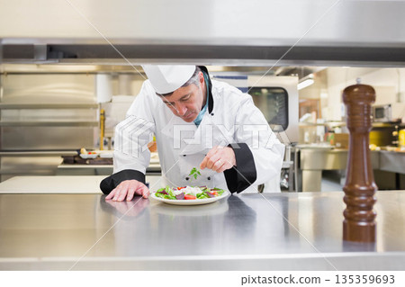 Male chef adding fresh herbs to salad on plate beside wooden pepper mill in professional kitchen Male chef adding fresh herbs to salad on plate beside wooden pepper mill in professional kitchen 135359693