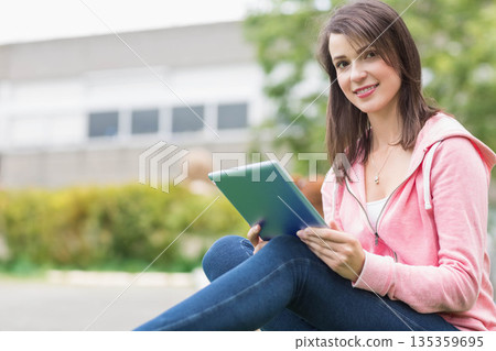 Woman sitting cross-legged on courtyard wearing pink hoodie holding tablet computer, copy space 135359695