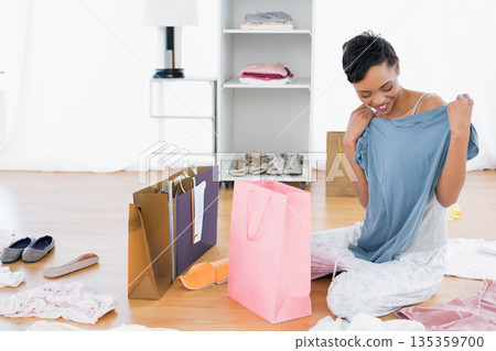 African American woman sitting in dressing room holding blue shirt with shopping bags, copy space 135359700