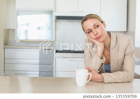 Woman wearing beige blazer leaning elbow on kitchen counter with coffee mug, copy space 135359709