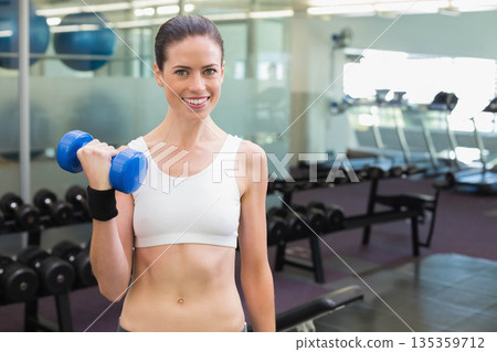 Woman in sportswear lifting blue dumbbell smiling at camera in gym with mirrored wall, treadmills Woman in sportswear lifting blue dumbbell smiling at camera in gym with mirrored wall, treadmills 135359712
