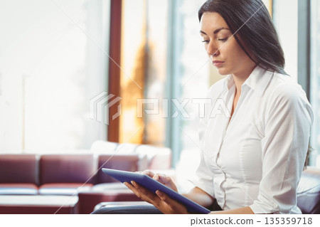 Businesswoman reviewing tablet content while sitting on cushioned sofa in office lounge, copy space 135359718