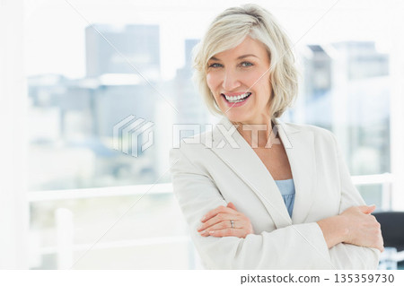 Middle-aged woman standing with arms crossed in office at window overlooking skyline, copy space Middle-aged woman standing with arms crossed in office at window overlooking skyline, copy space 135359730