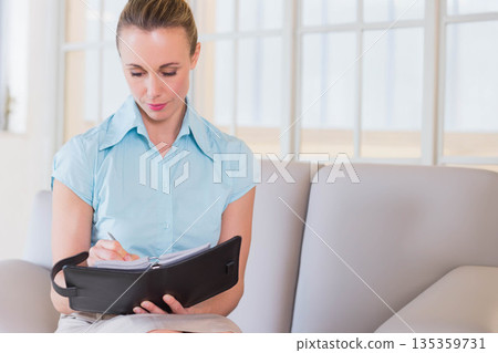Mid adult woman sitting on grey sofa in office lounge writing in black planner with pen 135359731