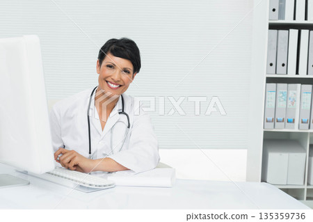 Female physician sitting at desk in office using monitor while wearing lab coat with stethoscope 135359736