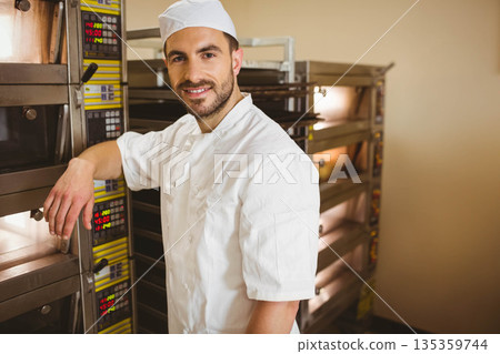 Male baker in uniform leaning on open oven door in bakery beside rack ovens, copy space 135359744