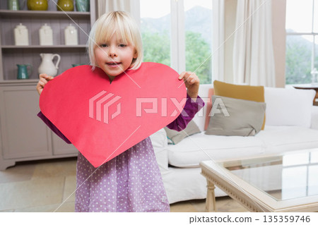 White female child standing by grey shelving unit in living room holding large red paper heart 135359746