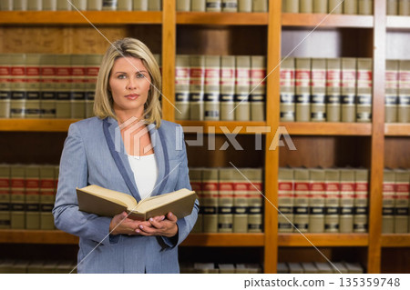 Woman wearing blue blazer reading open law book in law library with shelves of bound volumes 135359748