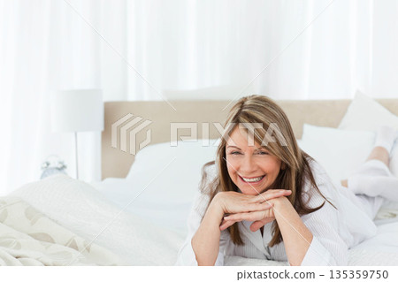Senior woman lying on bed in bedroom resting chin on hands smiling through sheer curtains 135359750
