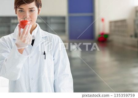 Female researcher wearing white lab coat, gloves inspecting red tomato in lab corridor, copy space Female researcher wearing white lab coat, gloves inspecting red tomato in lab corridor, copy space 135359759