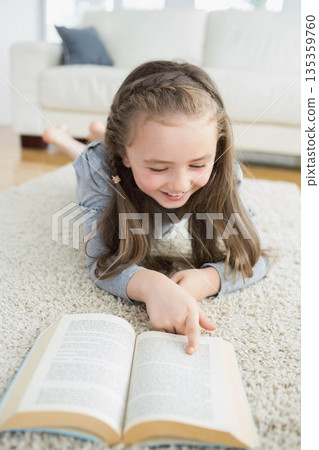 Girl child reading open book while lying on plush rug in living room with white sofa Girl child reading open book while lying on plush rug in living room with white sofa 135359760