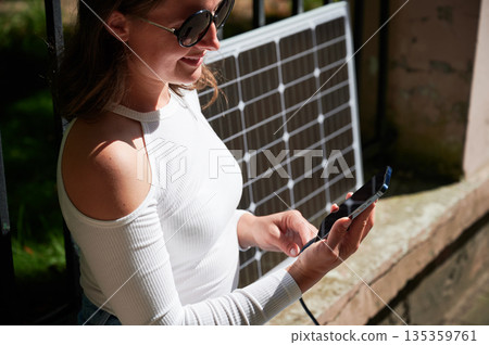 Woman connecting smartphone to photovoltaic solar panel. Cropped view. Integration of sustainable renewable energy into everyday life, demonstrating practical use of solar power for charging devices. Woman connecting smartphone to photovoltaic solar panel. Cropped view. Integration of sustainable renewable energy into everyday life, demonstrating practical use of solar power for charging devices. 135359761