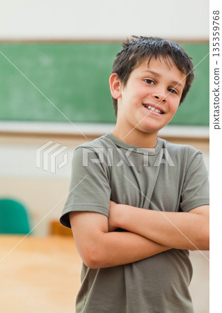 School-aged boy standing with arms crossed in classroom near wood desk, green chair and chalkboard School-aged boy standing with arms crossed in classroom near wood desk, green chair and chalkboard 135359768