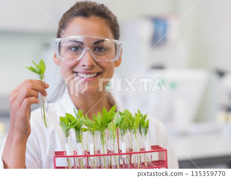 Female scientist wearing lab coat, safety goggles holding test tube with plant cutting in lab rack 135359770