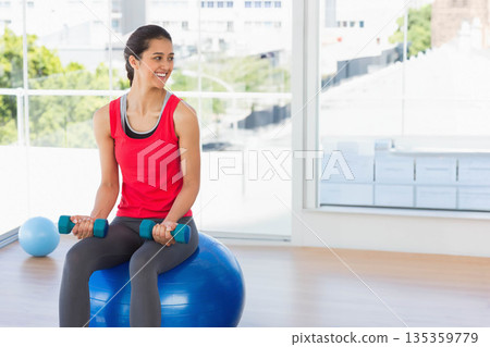 Woman in red top sitting on blue exercise ball in fitness studio, holding teal dumbbells 135359779