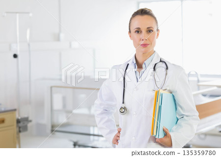 Woman doctor standing beside hospital bed wearing stethoscope and holding folders, copy space 135359780