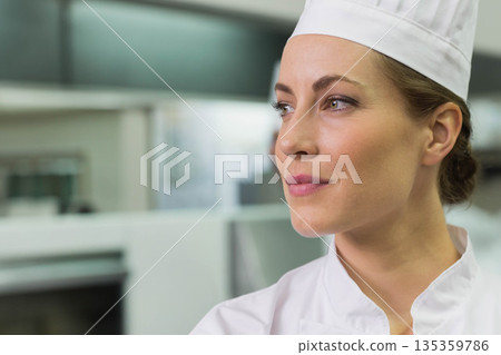 Mid adult female chef standing in kitchen by stainless counters wearing toque and coat, copy space 135359786