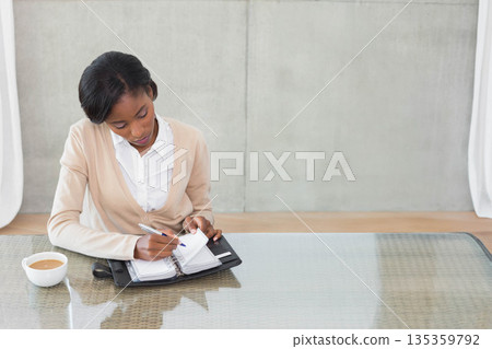 African American woman sitting at glass table writing planner with pen near coffee cup, copy space African American woman sitting at glass table writing planner with pen near coffee cup, copy space 135359792