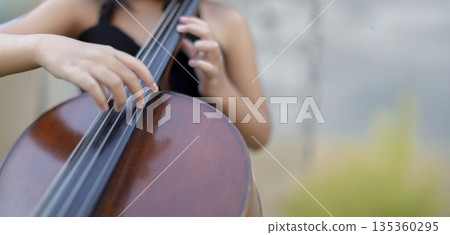 Close-up of a musician's hands playing cello in a green garden. Concept of classical music performance, outdoor practice, musical education, and nature-inspired melody 135360295