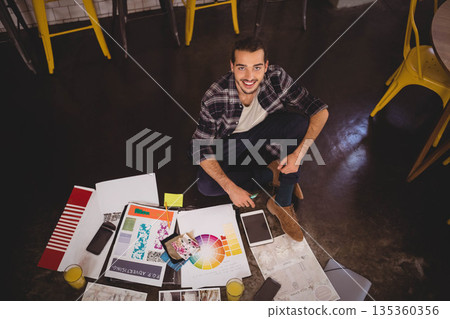 Man sitting cross legged on floor in design studio using smartphone amid design prints and juice Man sitting cross legged on floor in design studio using smartphone amid design prints and juice 135360356