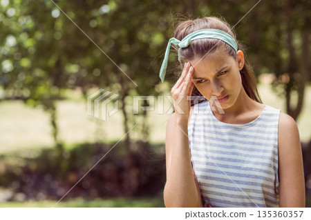 Teenage girl wearing headband and striped top holding temple furrowing brow in garden, copy space Teenage girl wearing headband and striped top holding temple furrowing brow in garden, copy space 135360357
