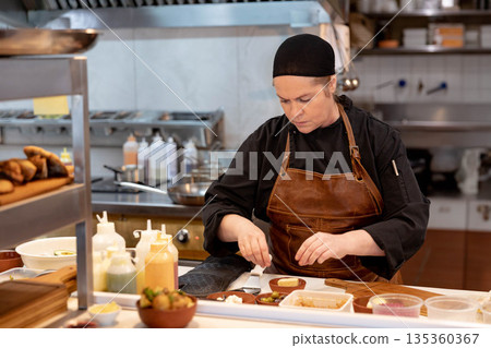 Female chef arranging small food items on plate on stainless steel counter in restaurant kitchen Female chef arranging small food items on plate on stainless steel counter in restaurant kitchen 135360367