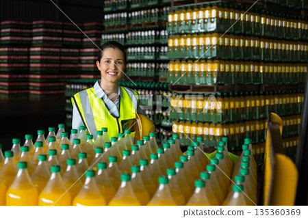 Female warehouse worker holding helmet wearing safety vest in warehouse among beverage bottles Female warehouse worker holding helmet wearing safety vest in warehouse among beverage bottles 135360369