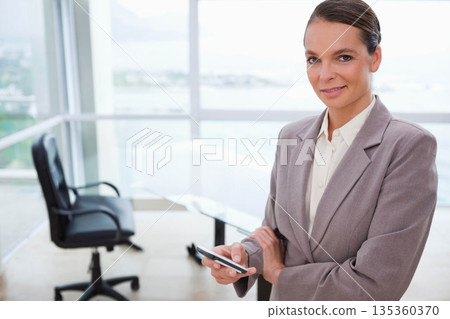 Female professional standing in glass office holding smartphone overlooking harbor, copy space 135360370