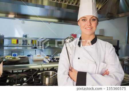Adult female chef standing by stainless steel stove holding ladle in restaurant kitchen, copy space Adult female chef standing by stainless steel stove holding ladle in restaurant kitchen, copy space 135360375