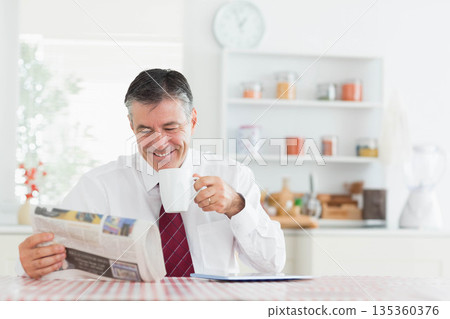 Middle-aged man wearing white shirt and tie reading newspaper holding coffee mug at kitchen table 135360376