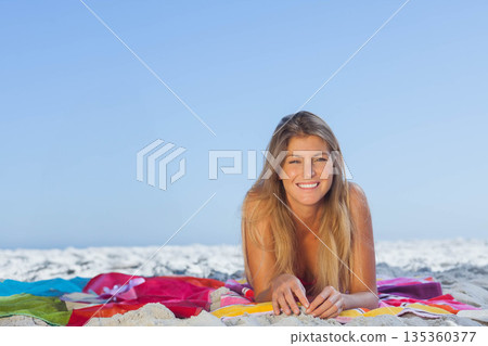 Woman lying on striped beach towel at seaside wearing red swimsuit, propping on elbows and smiling 135360377