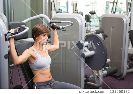 Woman wearing sports bra leggings using shoulder press machine in gym with mirrors, copy space Woman wearing sports bra leggings using shoulder press machine in gym with mirrors, copy space 135360382