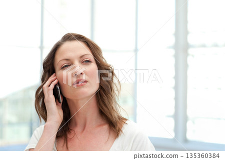 Mid-30s woman standing at modern office lobby holding smartphone to ear while looking right Mid-30s woman standing at modern office lobby holding smartphone to ear while looking right 135360384