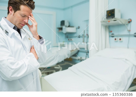 Mid-adult male doctor holding forehead in hospital ward with stethoscope and empty beds, copy space Mid-adult male doctor holding forehead in hospital ward with stethoscope and empty beds, copy space 135360386