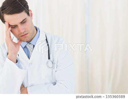 Male doctor pressing temple while wearing stethoscope by privacy curtains in exam room, copy space Male doctor pressing temple while wearing stethoscope by privacy curtains in exam room, copy space 135360389