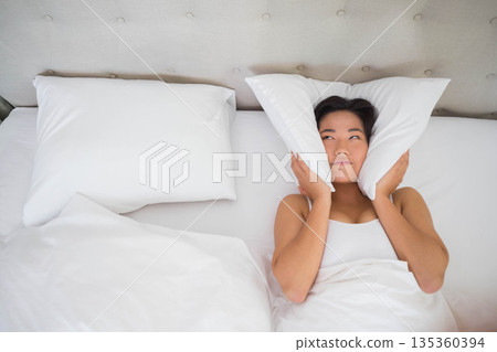 Korean woman lying in bed pressing pillows against ears under white duvet and headboard, copy space Korean woman lying in bed pressing pillows against ears under white duvet and headboard, copy space 135360394