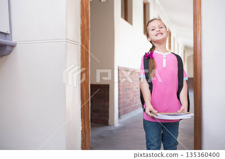 Female child standing in wooden door frame in school corridor holding notebooks and backpack 135360400