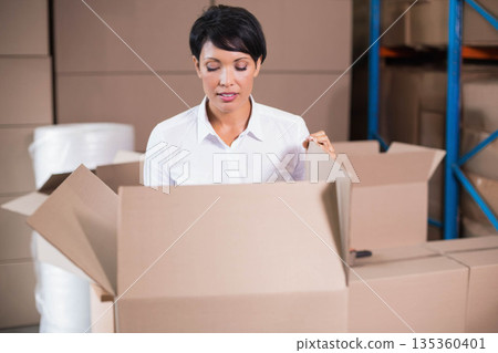 Woman in white shirt opening cardboard box in warehouse storage by bubble wrap and shelving rack 135360401