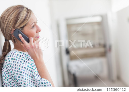 Woman standing in hospital corridor wearing gown and talking on smartphone near gurney, copy space 135360412