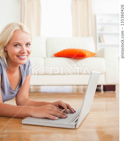 Light-haired woman lying in living room using silver laptop by sofa with orange cushion, copy space 135360426