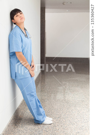 Hispanic woman nurse leaning on wall in hospital hallway wearing scrubs, stethoscope, copy space 135360427