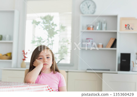 Child girl sitting at checkered table in kitchen under wall clock, gazing outside, copy space Child girl sitting at checkered table in kitchen under wall clock, gazing outside, copy space 135360429
