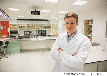 Male researcher wearing lab coat with arms crossed at medical lab near monitors and anatomy models 135360439