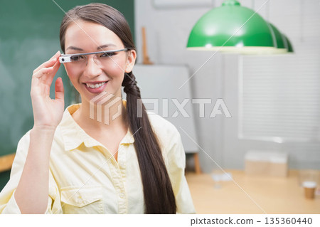 Asian female educator standing in classroom adjusting wearable smart glasses in front of chalkboard Asian female educator standing in classroom adjusting wearable smart glasses in front of chalkboard 135360440