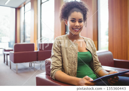 African American woman sitting in red leather armchair holding tablet computer near tall windows 135360443