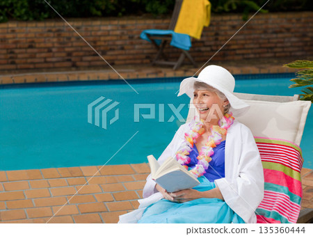 Senior woman wearing sun hat and floral lei reading book on pool patio chair, copy space Senior woman wearing sun hat and floral lei reading book on pool patio chair, copy space 135360444