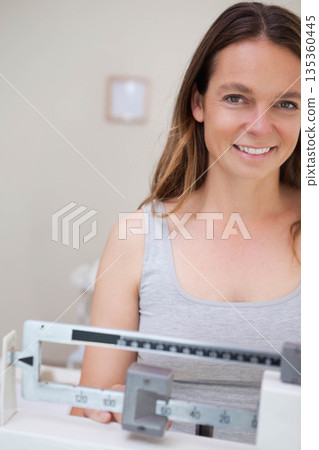 Woman standing behind mechanical beam scale in medical examination room wearing grey tank top Woman standing behind mechanical beam scale in medical examination room wearing grey tank top 135360445