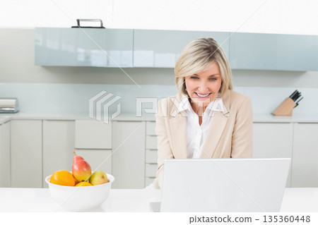 Woman in beige blazer typing on laptop at kitchen counter with fruit bowl and knife block Woman in beige blazer typing on laptop at kitchen counter with fruit bowl and knife block 135360448