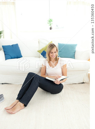 Woman sitting on carpet reading open book in living room by sofa with slippers and plant Woman sitting on carpet reading open book in living room by sofa with slippers and plant 135360450
