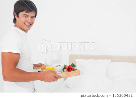 Man standing beside bed holding wooden tray with juice, cereal and rose in bedroom, copy space 135360451